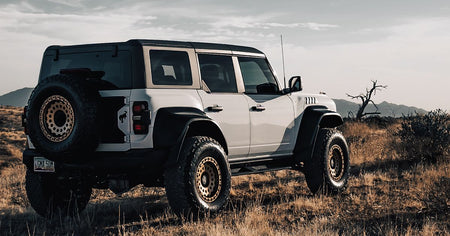 A white off-road SUV with a black soft top and large off-road tires parked in a dry grassy field with hills in the background.