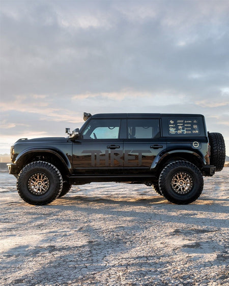 A black lifted Ford Bronco with oversized off-road tires and custom wheels, parked on a snowy, rocky ground under a cloudy sky.