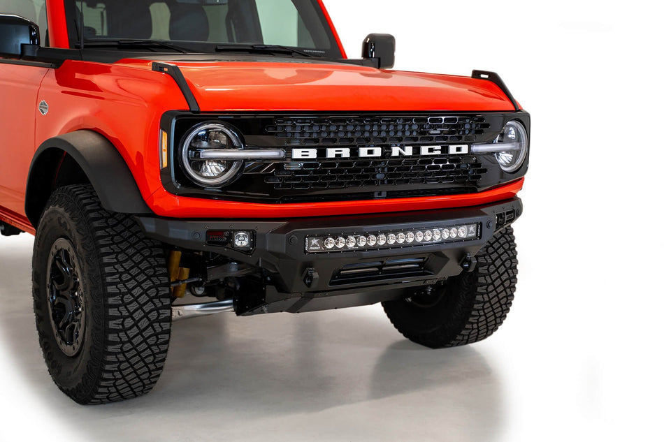 Addictive Desert Designs Stealth Fighter front bumper installed on orange Ford Bronco, close-up front three-quarter view showing grille and light bar