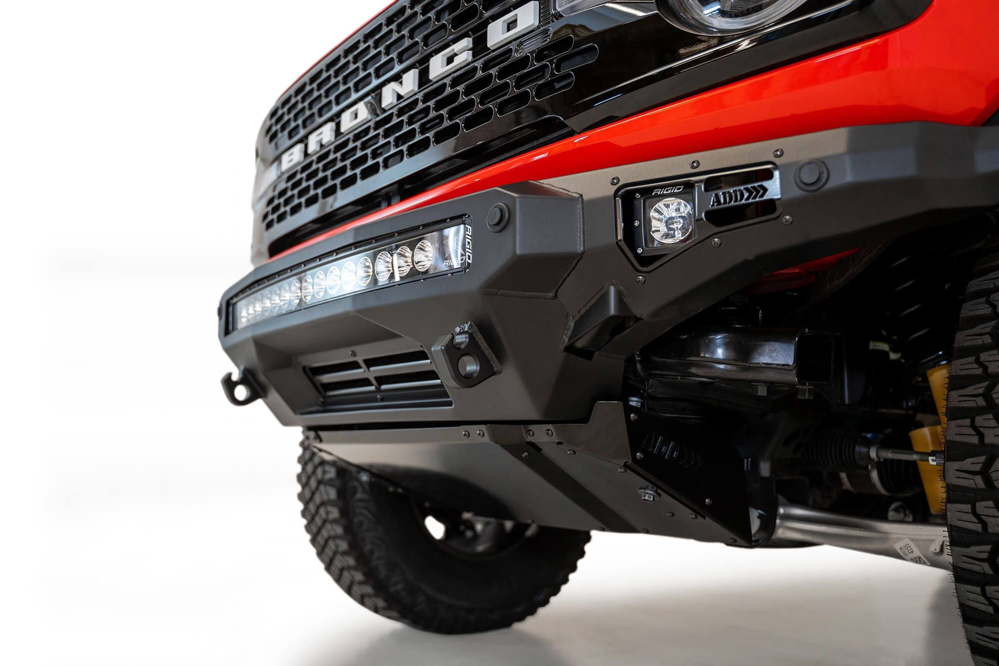 Addictive Desert Designs Stealth Fighter front bumper installed on orange Ford Bronco, close-up front three-quarter view product shot on white background