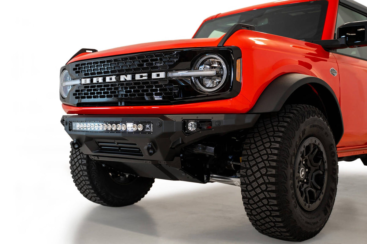 Addictive Desert Designs Stealth Fighter front bumper installed on orange Ford Bronco, front three-quarter close-up showing wheel and light bar, product shot on white background