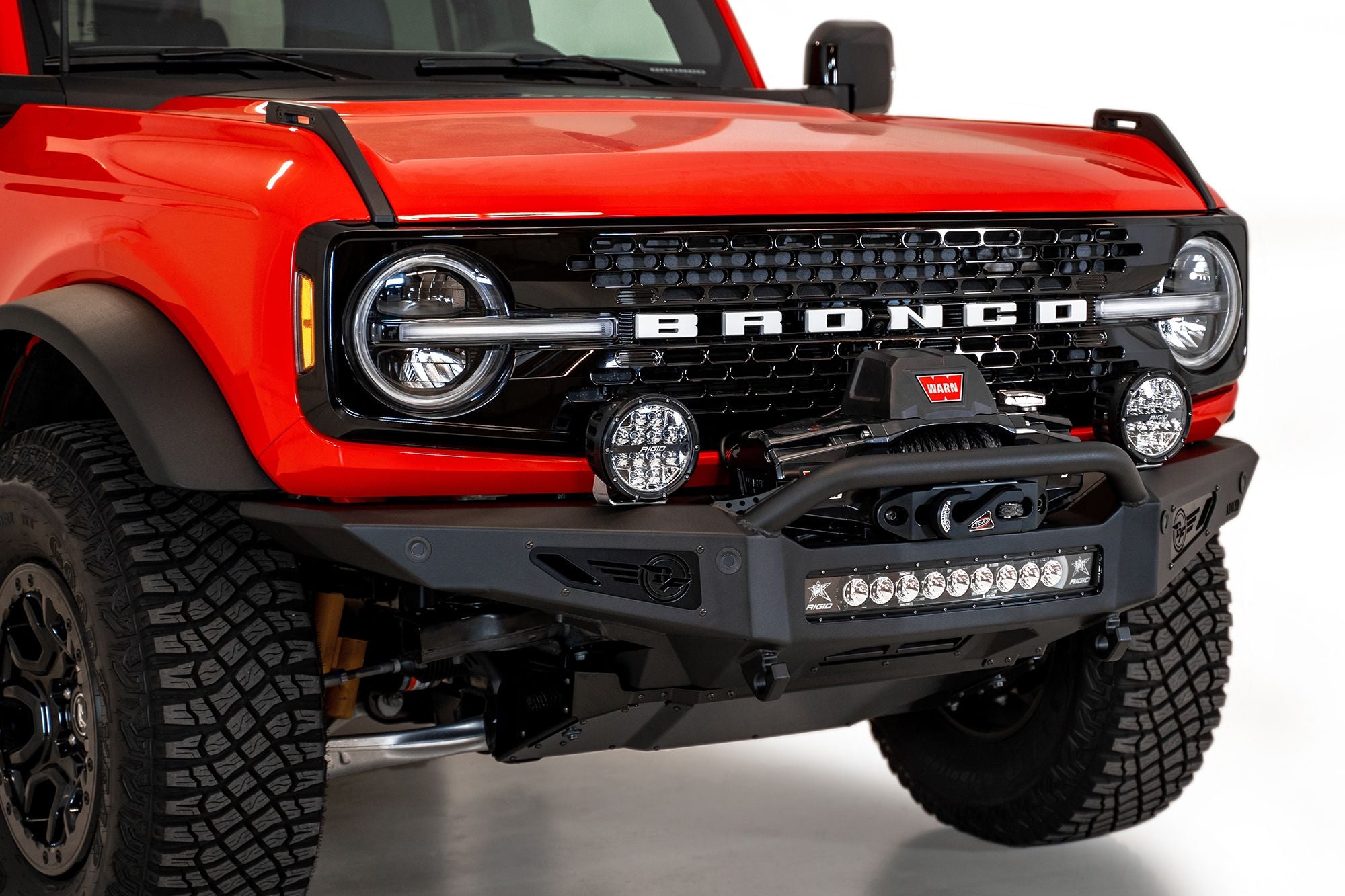 Addictive Desert Designs Rock Fighter winch front bumper installed on red Ford Bronco, close-up front three-quarter view showing winch and light bar