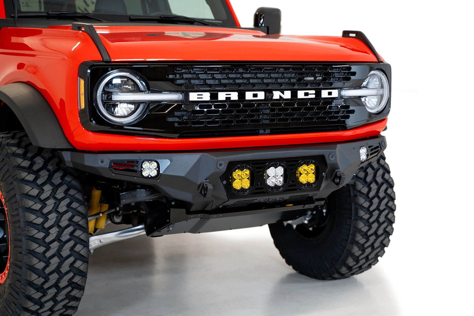 Addictive Desert Designs Bomber Baja front bumper installed on orange Ford Bronco, close-up front three-quarter view showing amber and white light pods