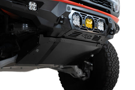 Addictive Desert Designs Bomber Baja front bumper installed on orange Ford Bronco Raptor, front three-quarter view showing full bumper width and light pods