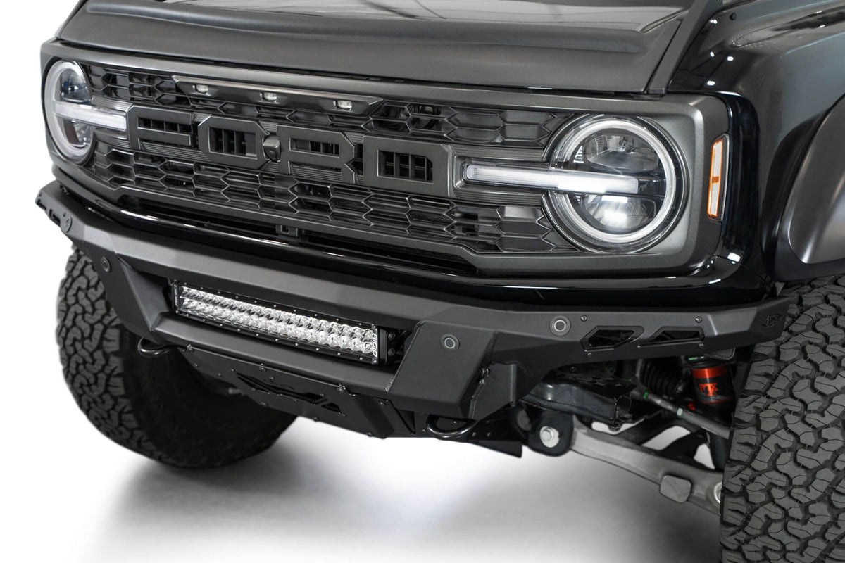 Addictive Desert Designs Phantom front bumper installed on black Ford Bronco Raptor, close-up showing grille, headlight, and integrated light bar