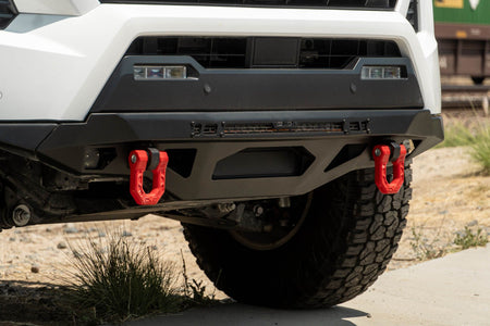 DV8 Offroad front bumper compatible with FBBO-01 fairlead cover plate, ground-level close-up showing red D-ring shackle and skid plate on gravel