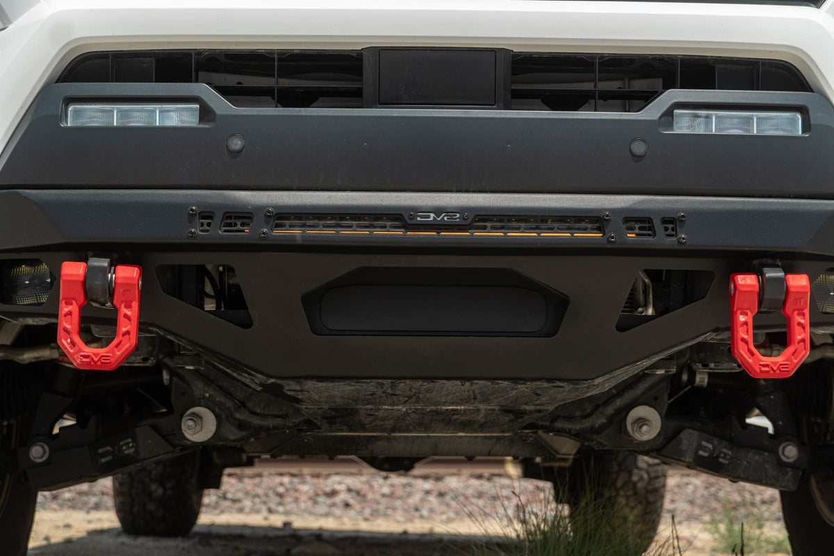 DV8 Offroad front bumper compatible with FBBO-01 fairlead cover plate, ground-level view showing skid plate and driver-side red recovery shackle