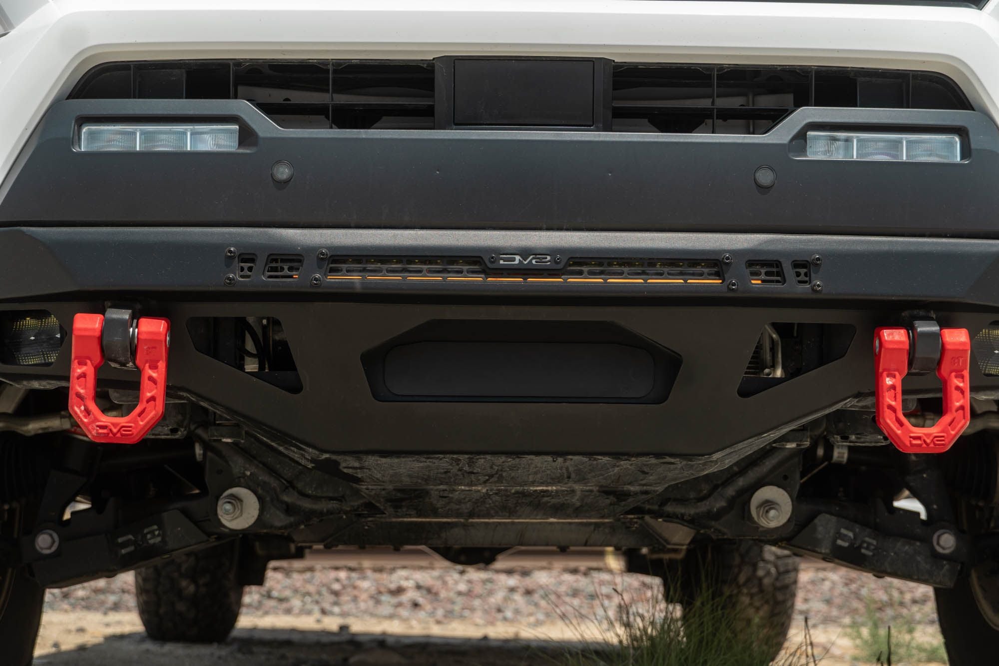 DV8 Offroad front bumper compatible with FBBO-01 fairlead cover plate, ground-level view showing skid plate and driver-side red recovery shackle