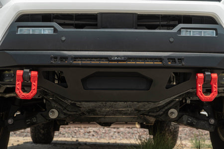 DV8 Offroad front bumper compatible with FBBO-01 fairlead cover plate, ground-level view showing skid plate and driver-side red recovery shackle