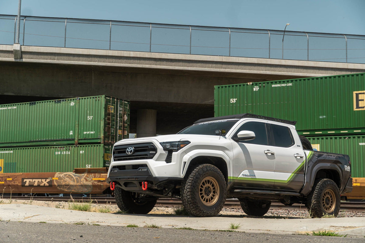 DV8 Offroad front bumper compatible with FBBO-01 fairlead cover plate, installed on Toyota Tacoma, wide front three-quarter view under highway overpass