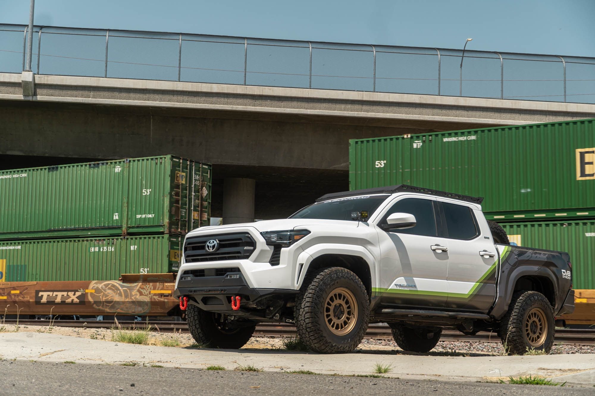 DV8 Offroad front bumper compatible with FBBO-01 fairlead cover plate, installed on Toyota Tacoma, wide front three-quarter view under highway overpass