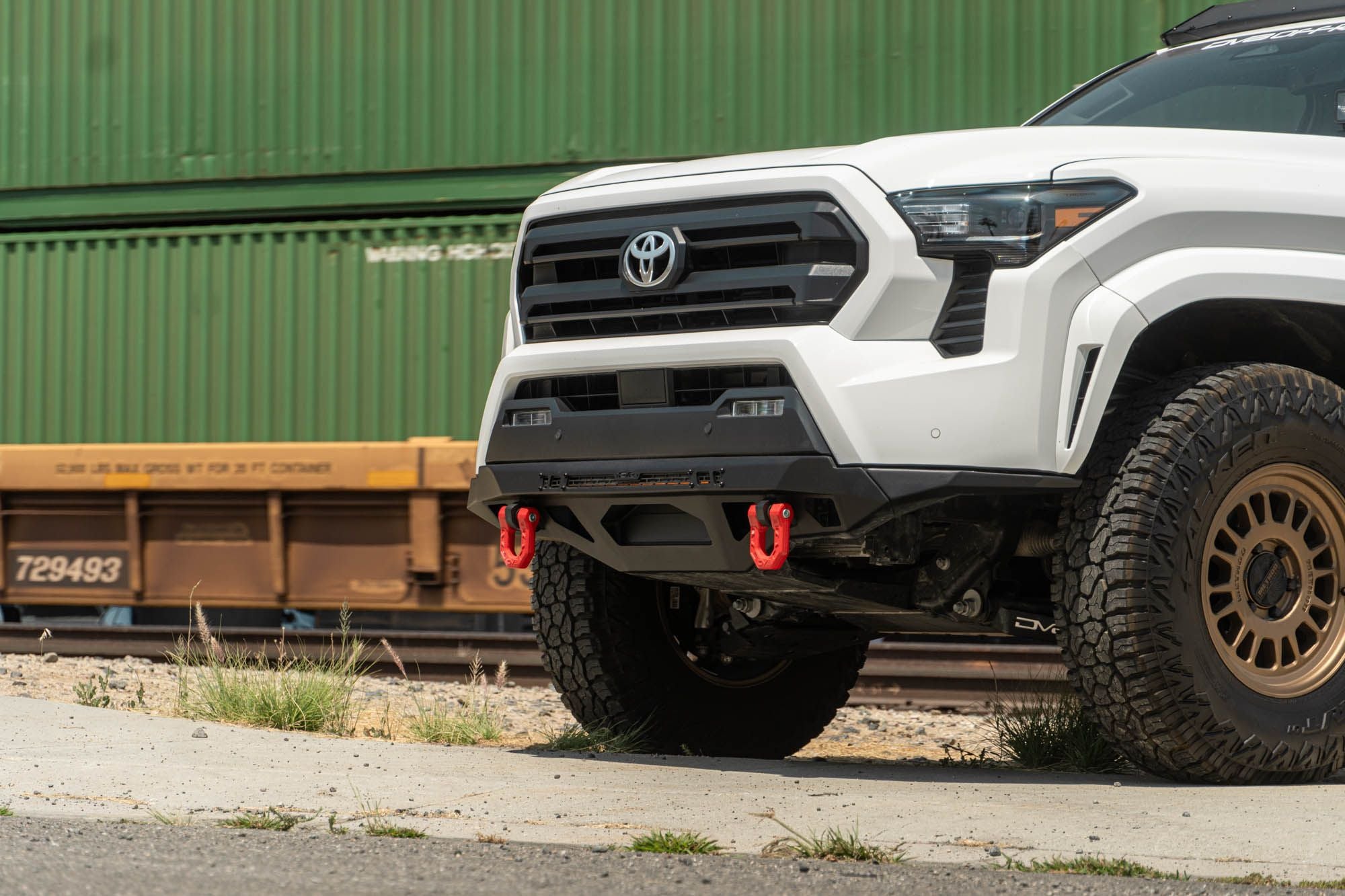 DV8 Offroad front bumper compatible with FBBO-01 fairlead cover plate, installed on Toyota Tacoma, front three-quarter view near shipping containers