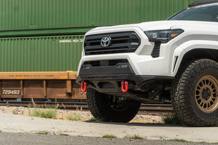 DV8 Offroad front bumper compatible with FBBO-01 fairlead cover plate, installed on Toyota Tacoma, front three-quarter view near shipping containers
