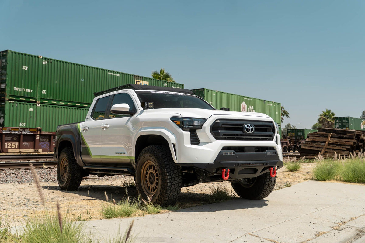 DV8 Offroad front bumper compatible with FBBO-01 fairlead cover plate, installed on Toyota Tacoma, wide front three-quarter view with shipping containers and blue sky