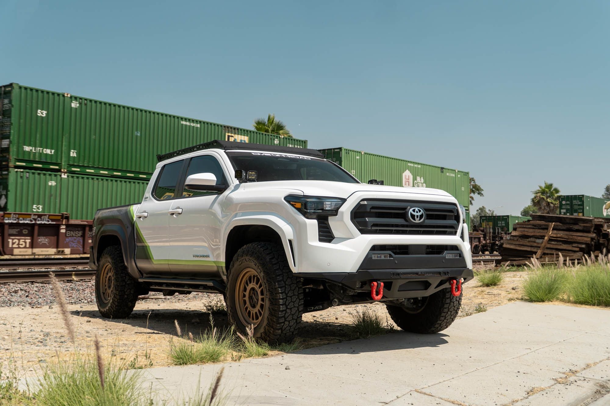 DV8 Offroad front bumper compatible with FBBO-01 fairlead cover plate, installed on Toyota Tacoma, wide front three-quarter view with shipping containers and blue sky