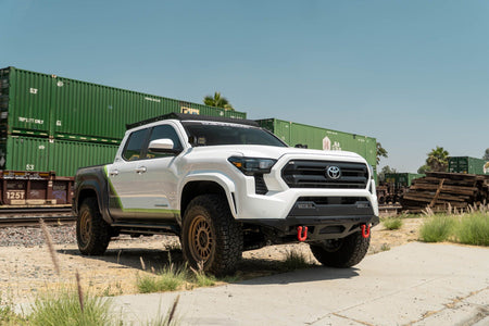 DV8 Offroad front bumper compatible with FBBO-01 fairlead cover plate, installed on Toyota Tacoma, wide front three-quarter view with shipping containers and blue sky