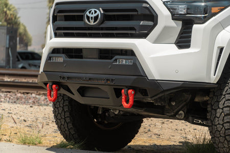DV8 Offroad front bumper compatible with FBBO-01 fairlead cover plate, installed on white Toyota Tacoma, front three-quarter view near railroad tracks