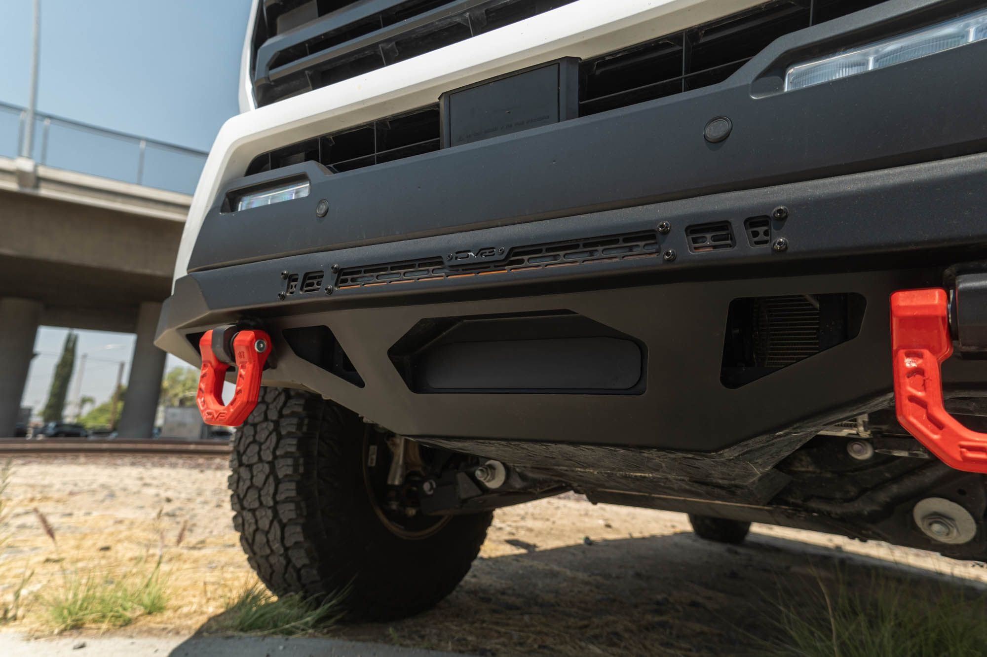DV8 Offroad front bumper compatible with FBBO-01 fairlead cover plate, low-angle view showing wheel, tire, red recovery shackle, and light bar under overpass