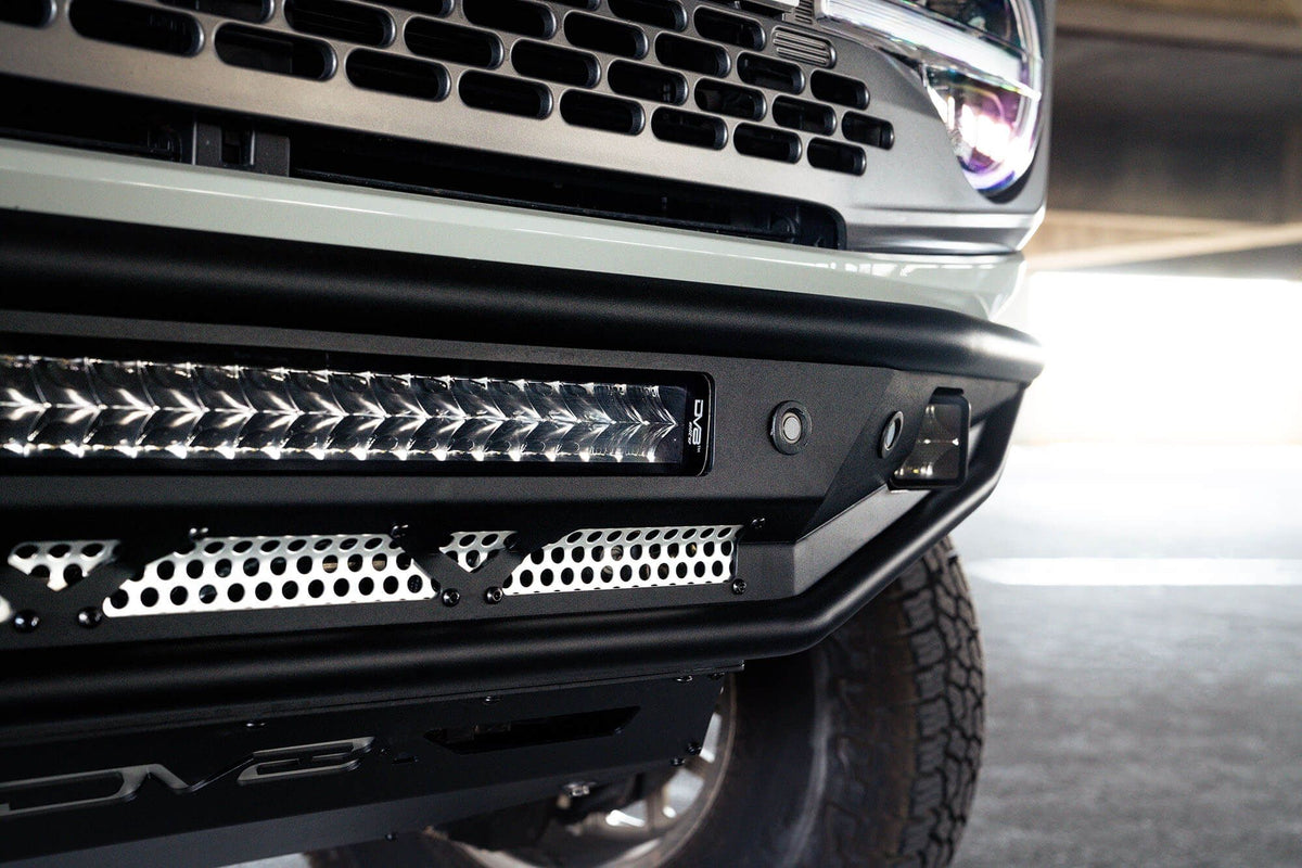 DV8 Offroad Competition Series front bumper installed on light-colored Ford Bronco, close-up showing grille and light bar in parking garage