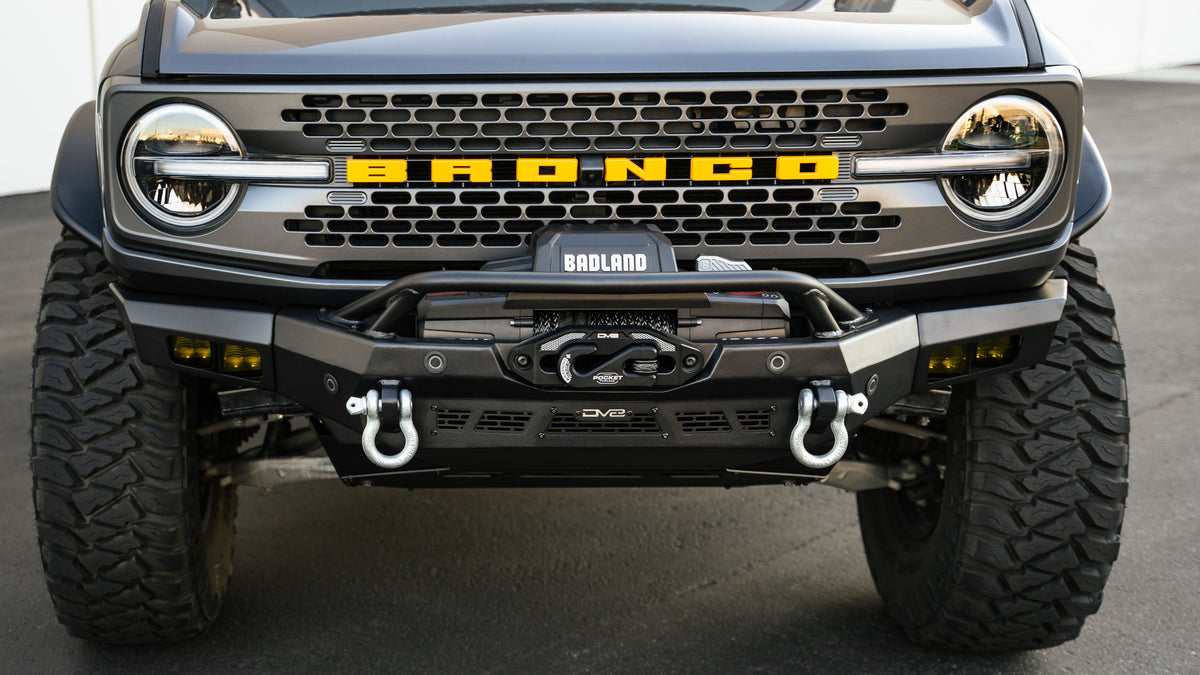 DV8 Offroad Spec Series winch front bumper installed on dark green Ford Bronco Badlands, straight-on front view showing D-ring shackles