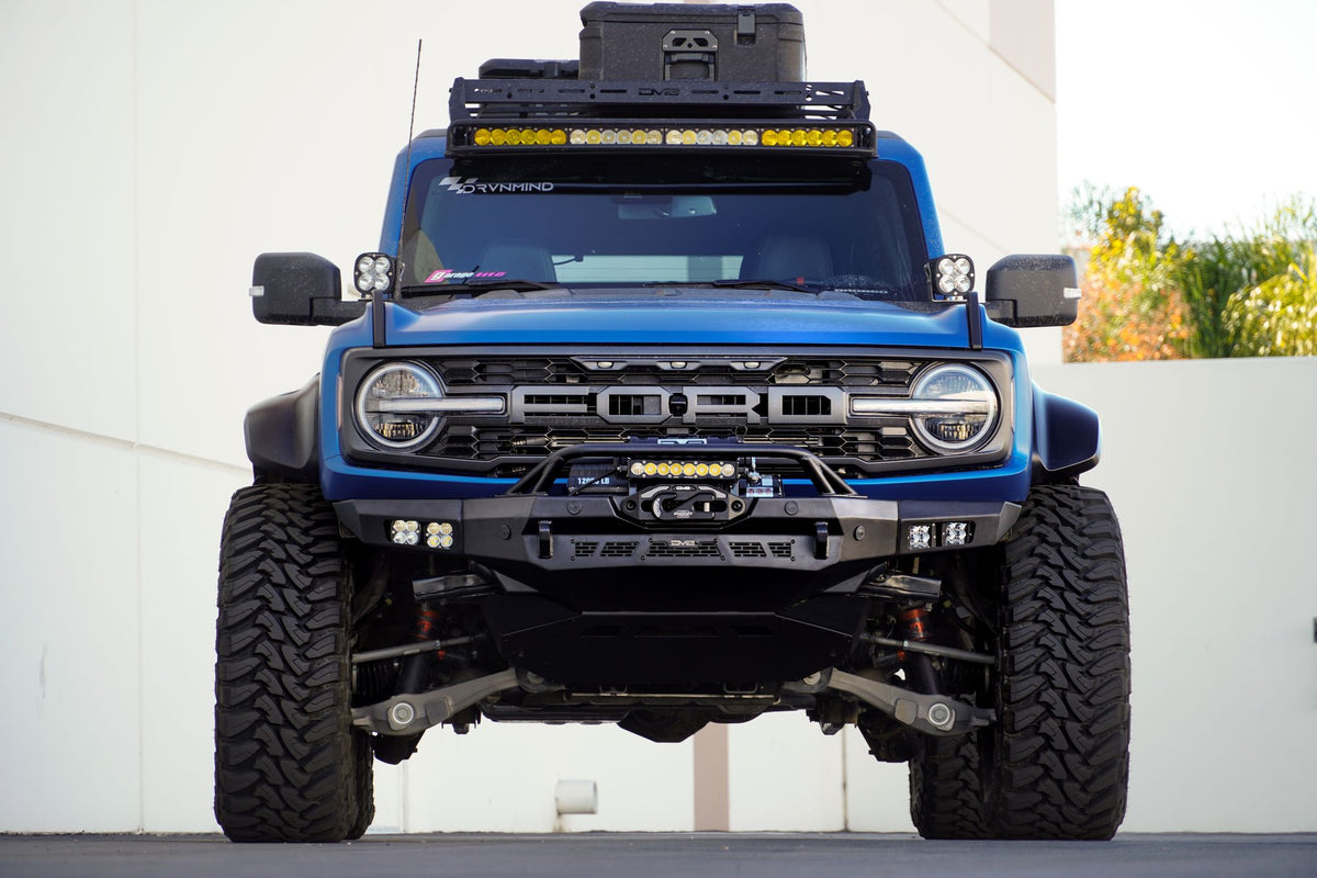 DV8 Offroad Spec Series winch front bumper installed on blue Ford Bronco with roof rack, straight-on front view in parking lot