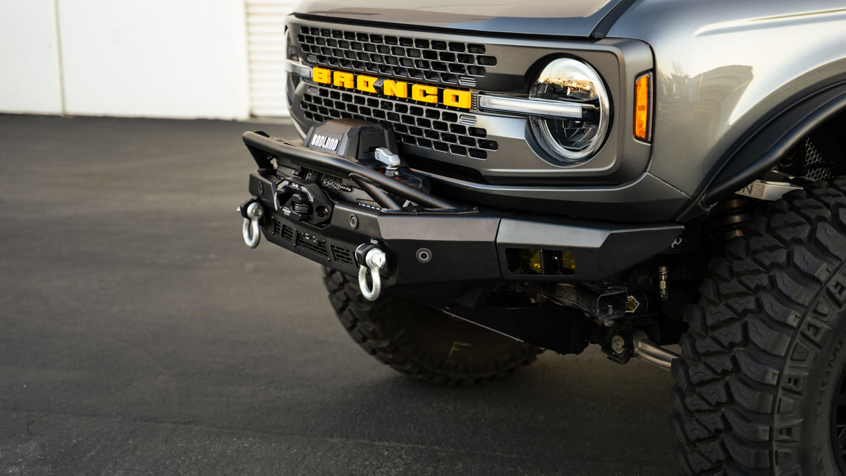 DV8 Offroad Spec Series winch front bumper installed on gray Ford Bronco, close-up front view showing BRONCO grille badge and shackles