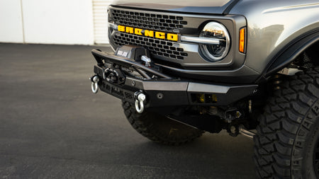 DV8 Offroad Spec Series winch front bumper installed on gray Ford Bronco, close-up front view showing BRONCO grille badge and shackles
