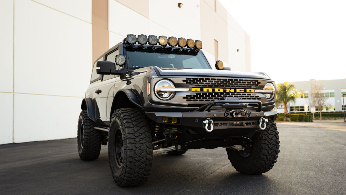 DV8 Offroad Spec Series winch front bumper installed on tan Ford Bronco with roof rack lights, distant front three-quarter view in parking lot