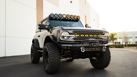 DV8 Offroad Spec Series winch front bumper installed on tan Ford Bronco with roof rack lights, distant front three-quarter view in parking lot
