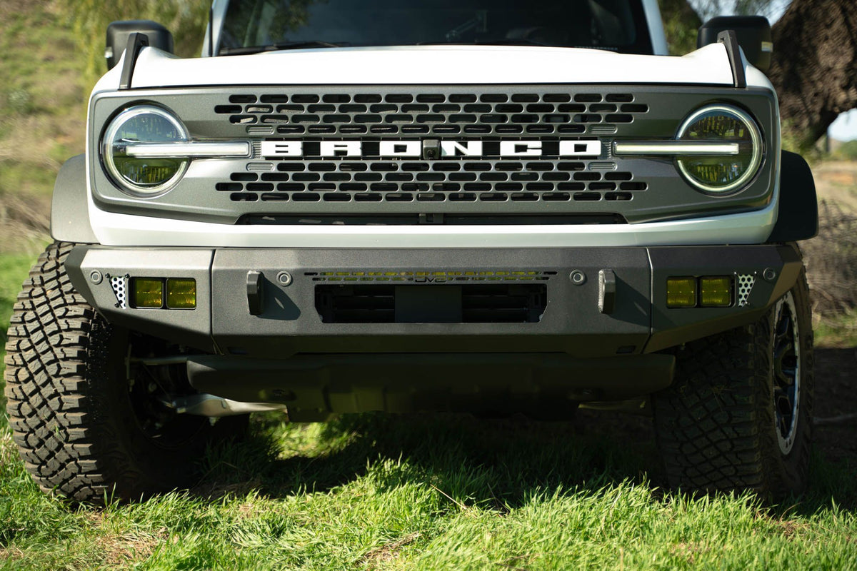DV8 Offroad OE Plus Series V2 front bumper installed on olive green Ford Bronco, close-up showing grille and bumper on hillside grass