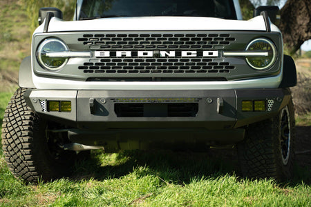 DV8 Offroad OE Plus Series V2 front bumper installed on olive green Ford Bronco, close-up showing grille and bumper on hillside grass