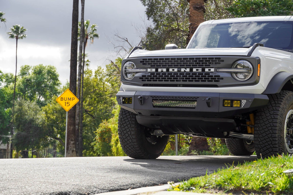 DV8 Offroad OE Plus Series V2 front bumper installed on white Ford Bronco, close-up front three-quarter view with palm trees and street sign