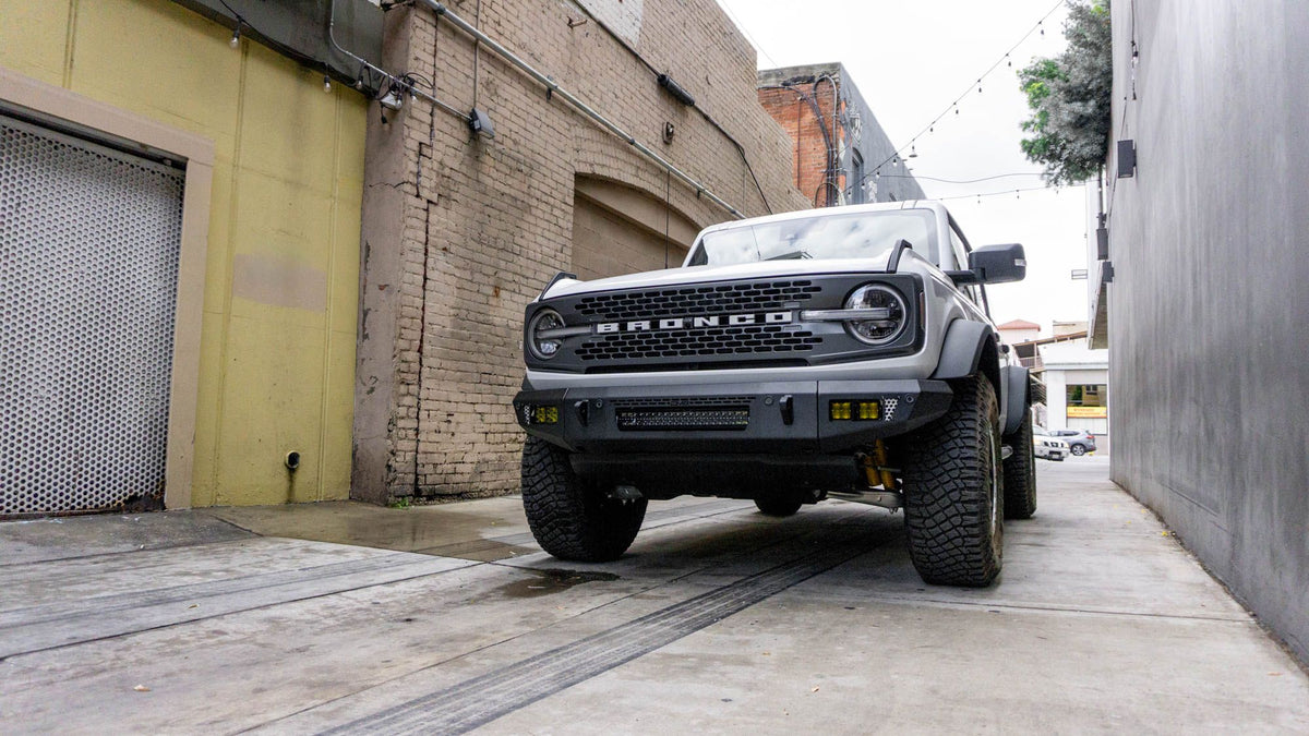 DV8 Offroad OE Plus Series V2 front bumper installed on white Ford Bronco, distant front three-quarter view near brick building