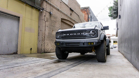 DV8 Offroad OE Plus Series V2 front bumper installed on white Ford Bronco, distant front three-quarter view near brick building