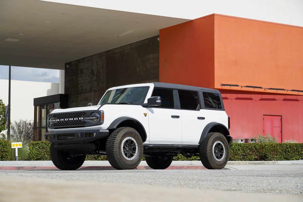 DV8 Offroad OE Plus Series V2 front bumper installed on white Ford Bronco, distant front three-quarter view near modern building