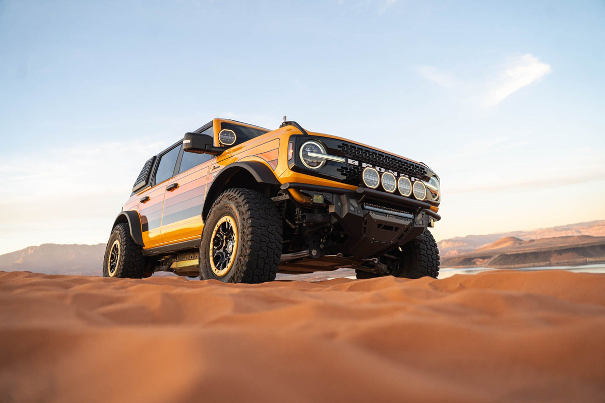 DV8 Offroad Tube Series front bumper installed on orange Ford Bronco, distant front three-quarter view on desert dunes