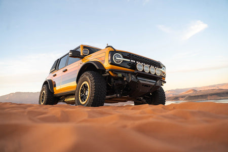 DV8 Offroad Tube Series front bumper installed on orange Ford Bronco, distant front three-quarter view on desert dunes