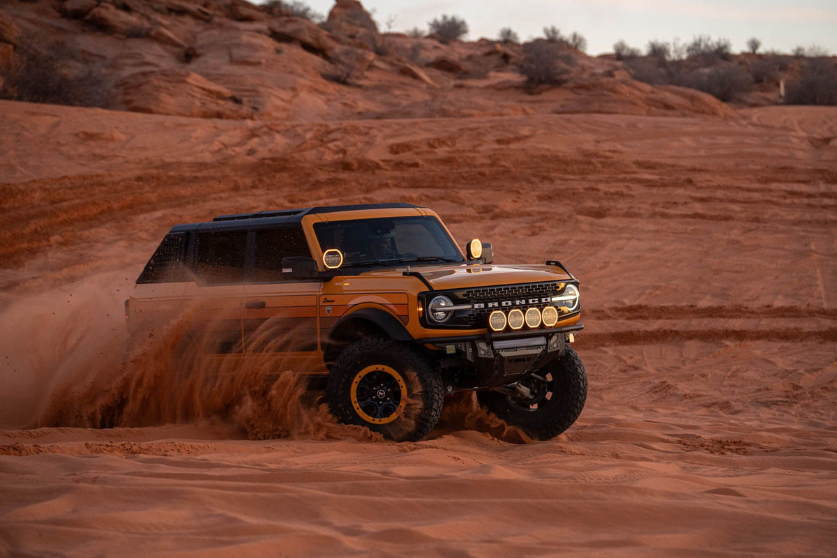 DV8 Offroad Tube Series front bumper installed on orange Ford Bronco, distant front three-quarter view kicking up dust on red rock desert