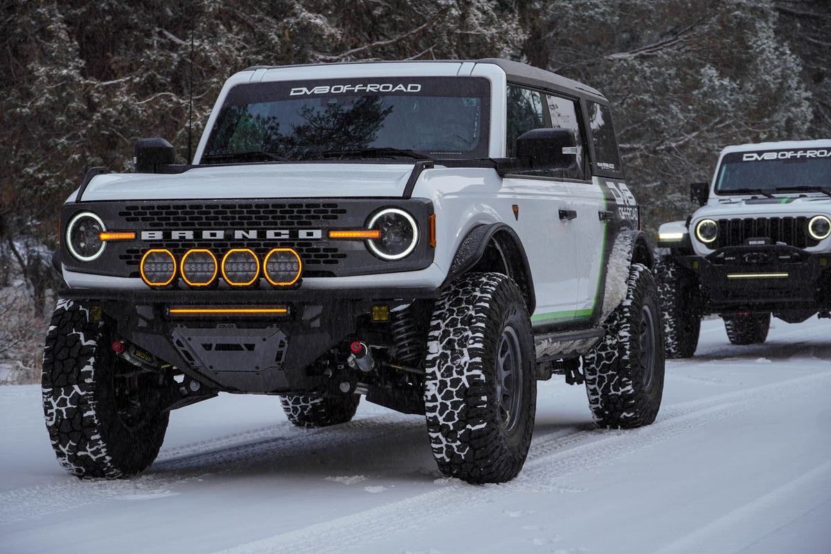 DV8 Offroad Tube Series front bumper installed on white Ford Bronco, front three-quarter view with second Bronco behind on snowy road