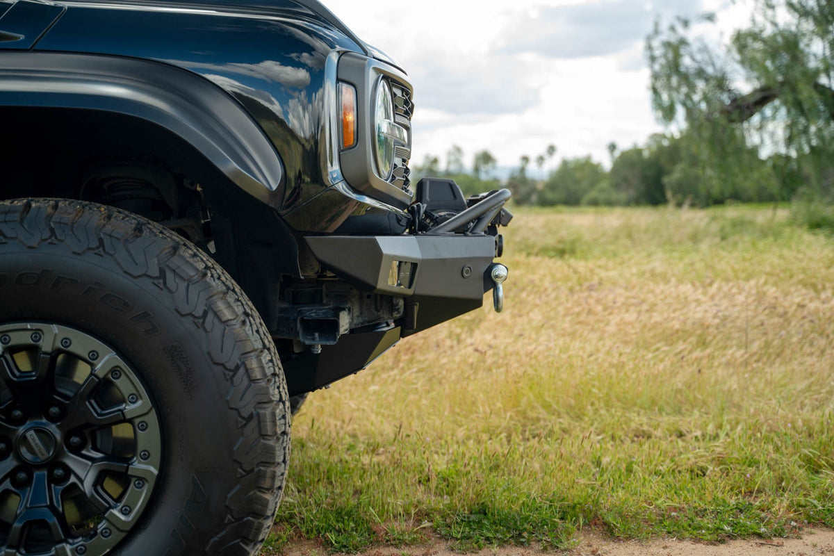 DV8 Offroad Spec Series winch front bumper installed on black Ford Bronco Raptor, close-up showing bumper corner and wheel in field
