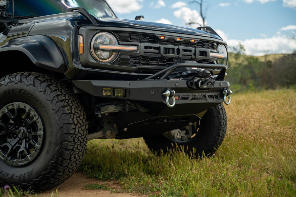 DV8 Offroad Spec Series winch front bumper installed on black Ford Bronco Raptor, close-up front three-quarter view in grass field