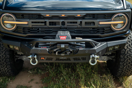 DV8 Offroad Spec Series winch front bumper installed on black Ford Bronco Raptor, straight-on close-up showing winch and D-ring shackles