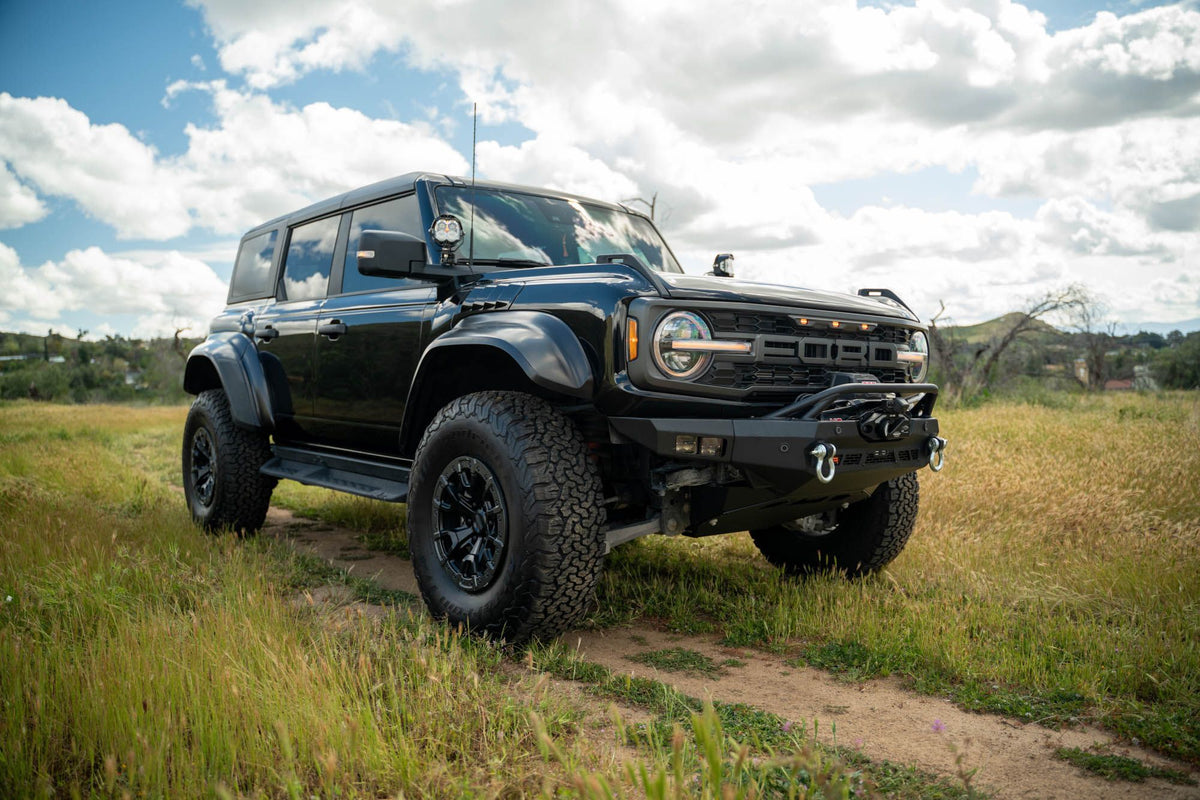 DV8 Offroad Spec Series winch front bumper installed on black Ford Bronco Raptor, distant front three-quarter view in field, alternate angle