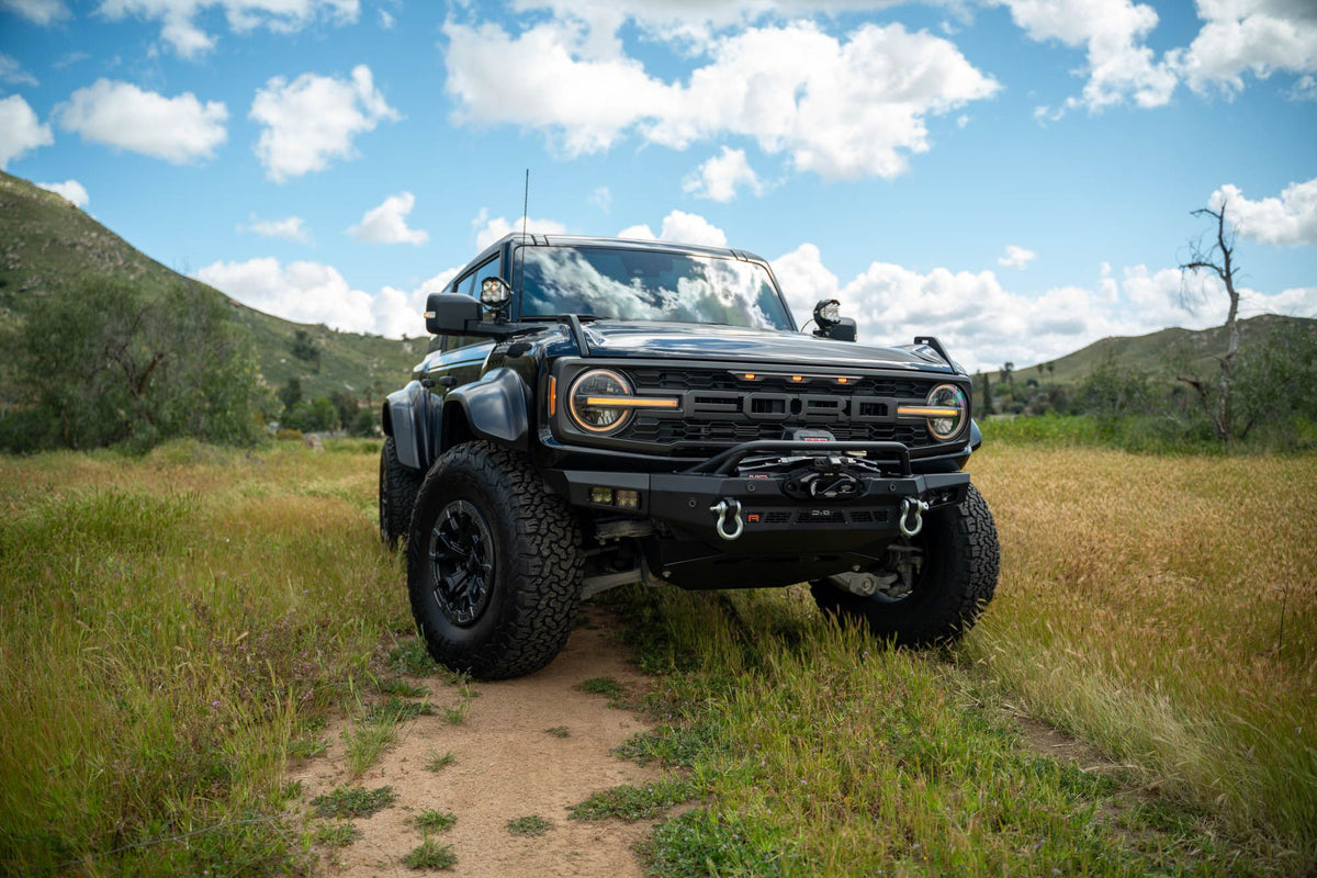 DV8 Offroad Spec Series winch front bumper installed on black Ford Bronco Raptor, distant front three-quarter view on mountain trail
