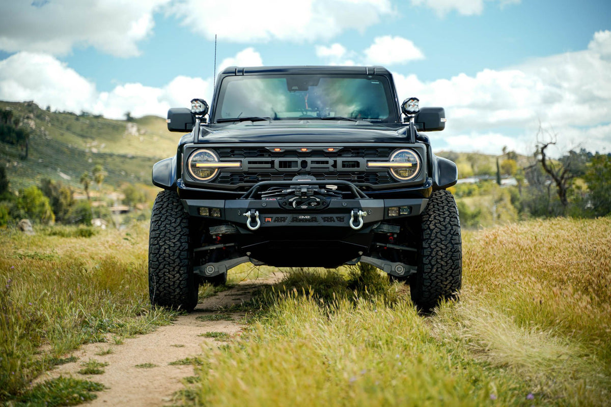 DV8 Offroad Spec Series winch front bumper installed on black Ford Bronco Raptor, straight-on front view on dirt trail in field