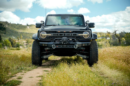 DV8 Offroad Spec Series winch front bumper installed on black Ford Bronco Raptor, straight-on front view on dirt trail in field