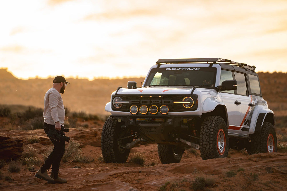 DV8 Offroad Tube Series front bumper installed on white Ford Bronco Raptor, distant front three-quarter view in desert at sunset with person standing nearby