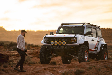 DV8 Offroad Tube Series front bumper installed on white Ford Bronco Raptor, distant front three-quarter view in desert at sunset with person standing nearby