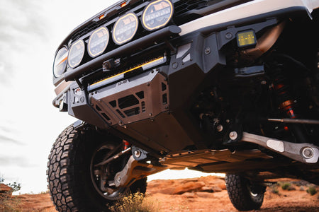 DV8 Offroad Tube Series front bumper installed on tan Ford Bronco Raptor, underside close-up on red rock desert terrain