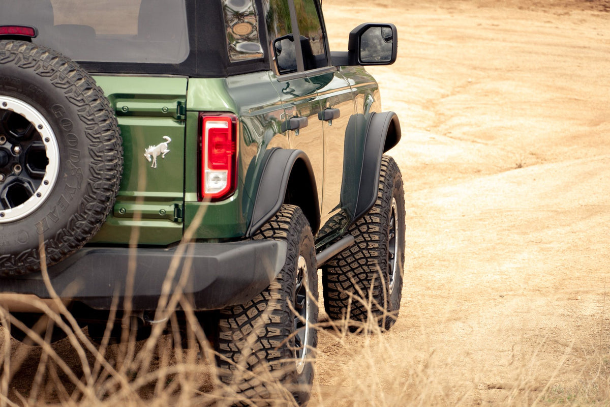 DV8 Offroad tube fender flares installed on green 4-door Ford Bronco, rear three-quarter view with spare tire on dirt field